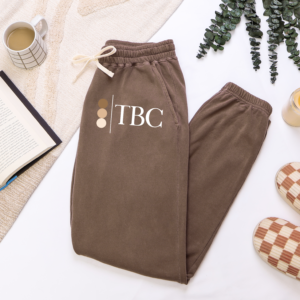 Folded Espresso brown joggers with The Bronze Collective brandmark logo on the leg, styled with a book, coffee, and slippers
