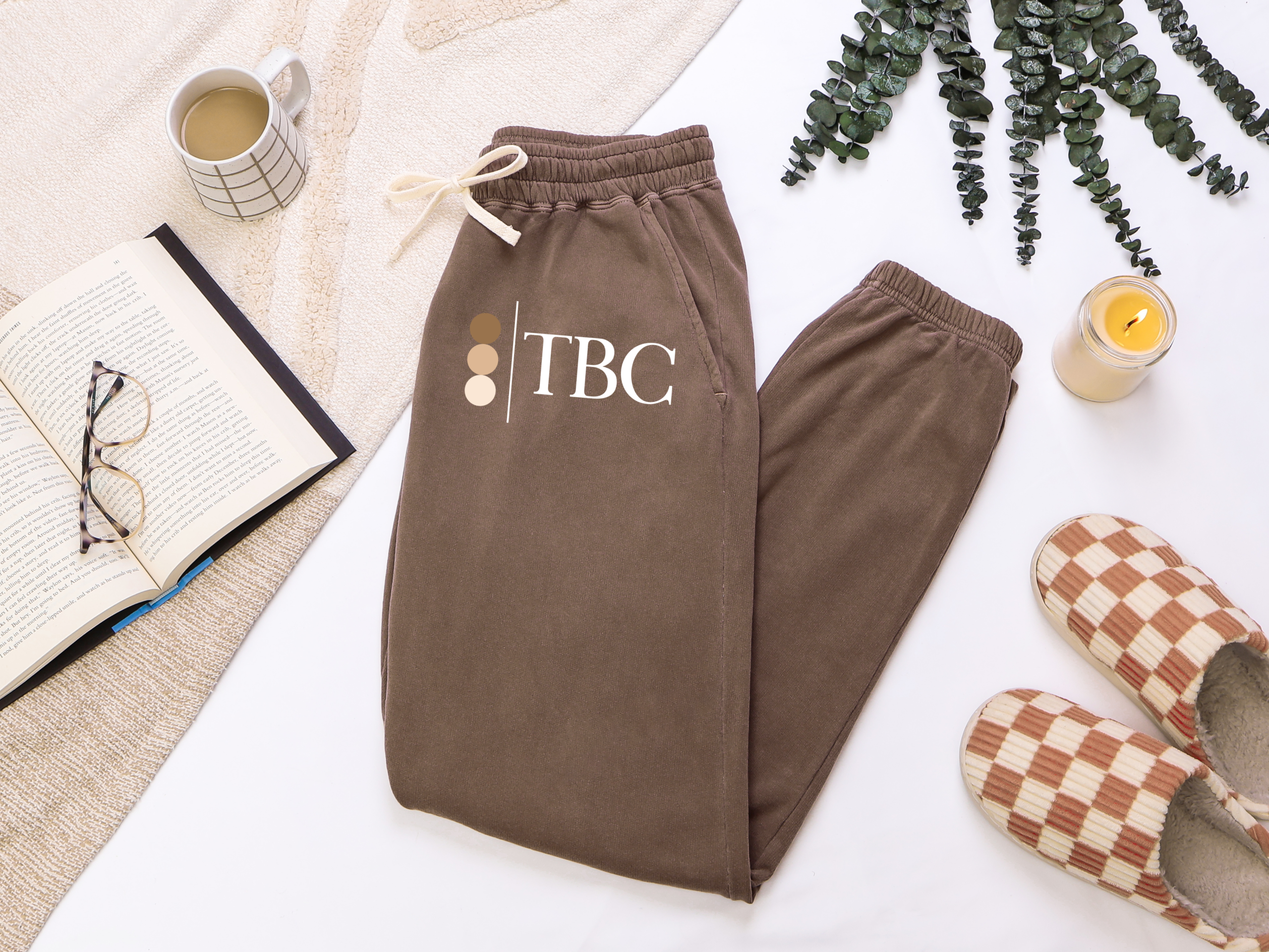 Folded Espresso brown joggers with The Bronze Collective brandmark logo on the leg, styled with a book, coffee, and slippers
