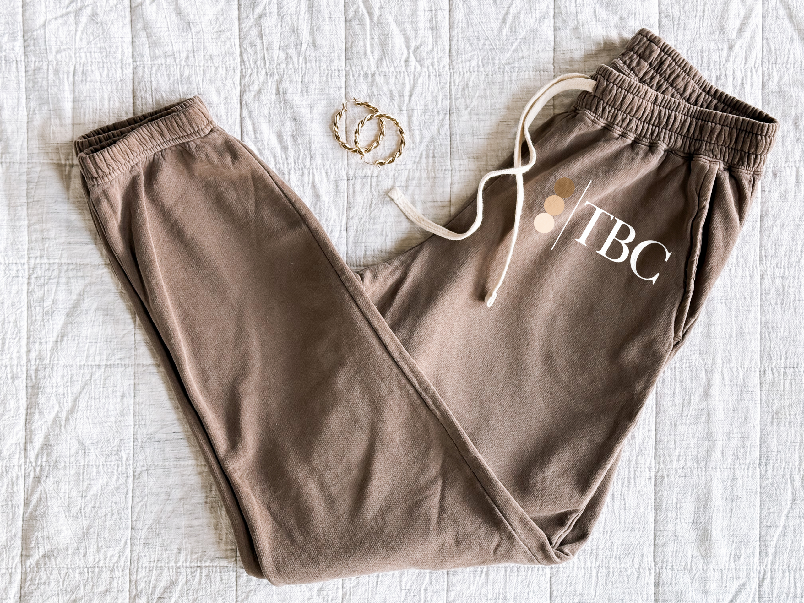 The Bronze Collective brandmark logo joggers in Espresso brown laid flat on a white quilt with gold hoop earrings